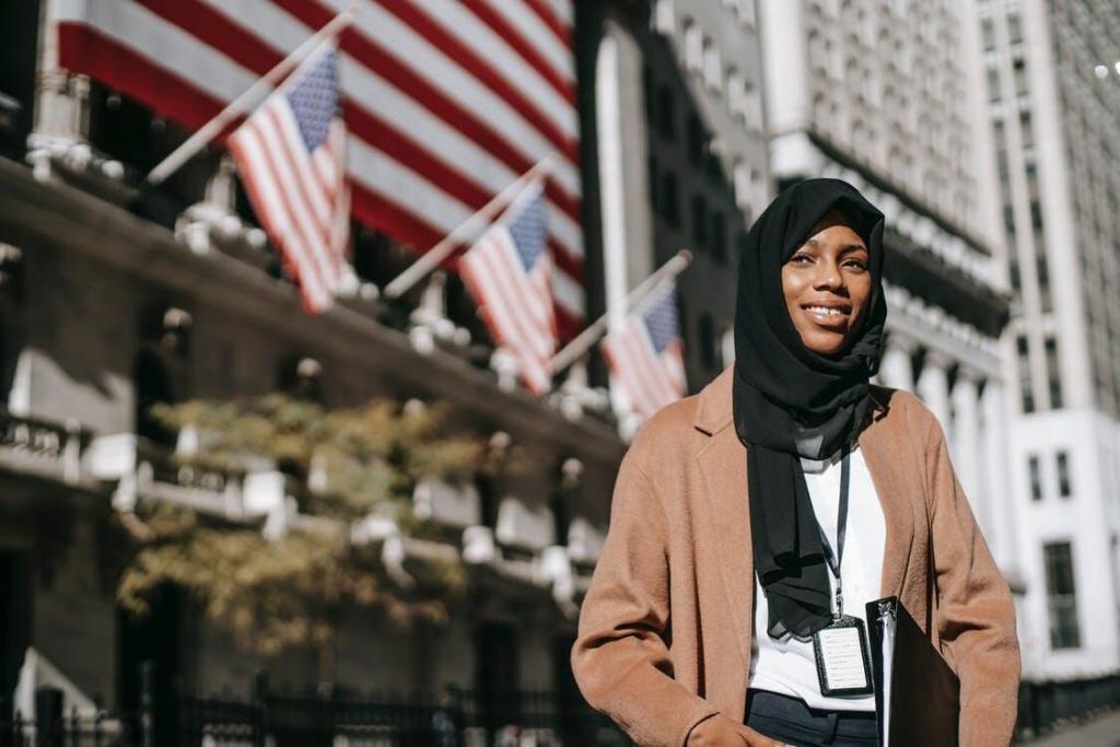 From below of cheerful African American female ambassador with folder wearing hijab and id card looking away while standing near building with American flags on blurred background
